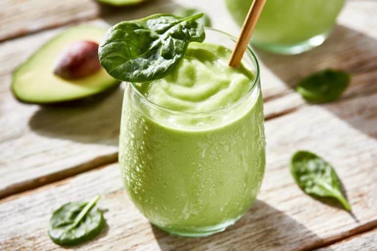 Un smoothie vert onctueux dans un verre givré avec une paille entouré d'avocats et de feuilles d'épinards frais
