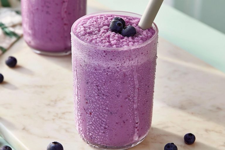 Un milkshake crémeux à la myrtille dans un verre givré entouré de quelques fruits frais sur une table marbrée